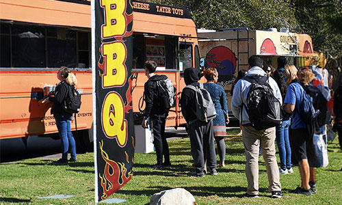 People lining up for food truck service next to a BBQ feather flag at an outdoor festival