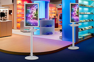 Freestanding digital kiosks with personal device charging line the aisle of a trade show floor.