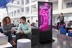 Groups of college students at a campus sit and chat next to an informational digital kiosk.