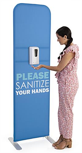 Woman sanitizing hands at the "Please Sanitize Your Hands" tension fabric banner with sanitizer dispenser