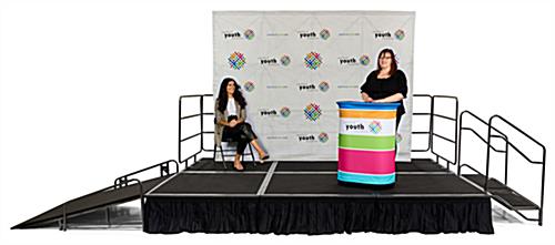 Front view of event stage with woman at a custom printed podium and another woman seated in front of a custom printed quick fabric backwall.